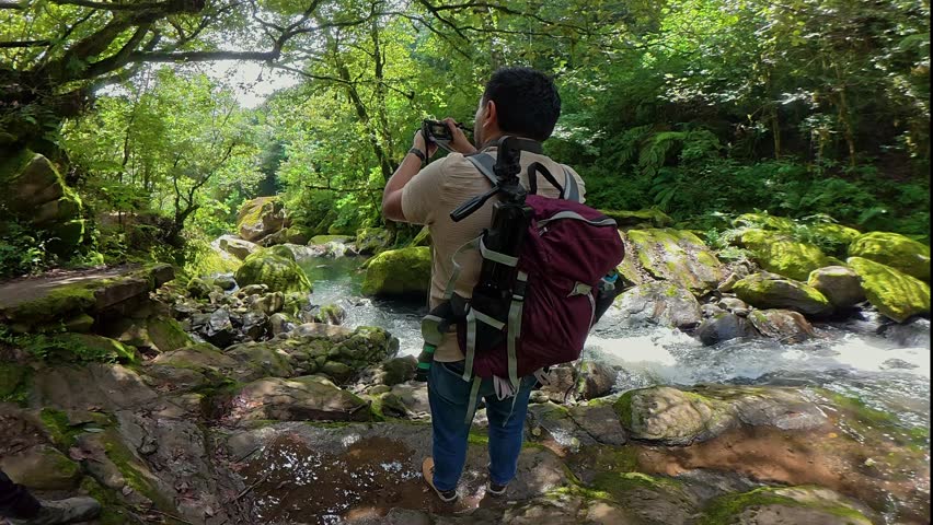 Male photographer with a backpack taking pictures of a river flowing through a lush green forest