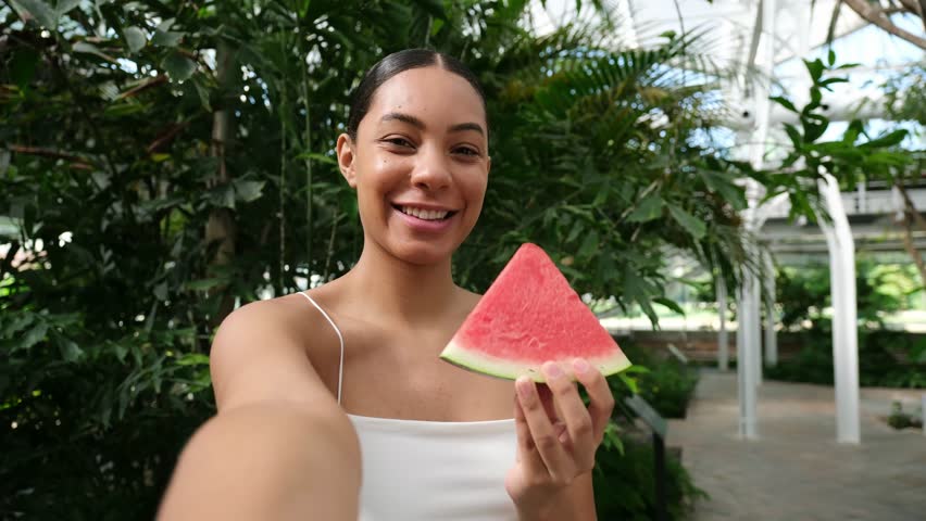 Young happy woman holding a slice of watermelon taking a selfie video in a botanical garden