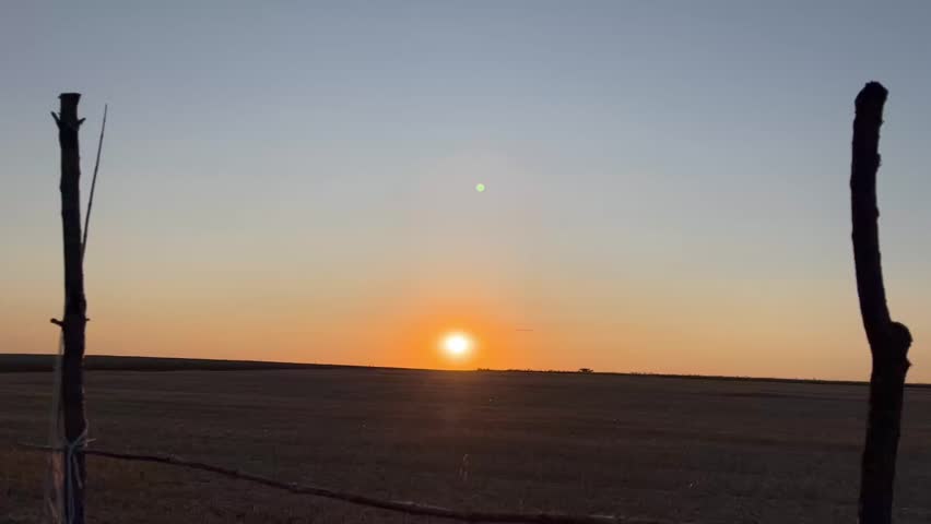 Bright sun setting over a wide open rural field with a wooden fence.
