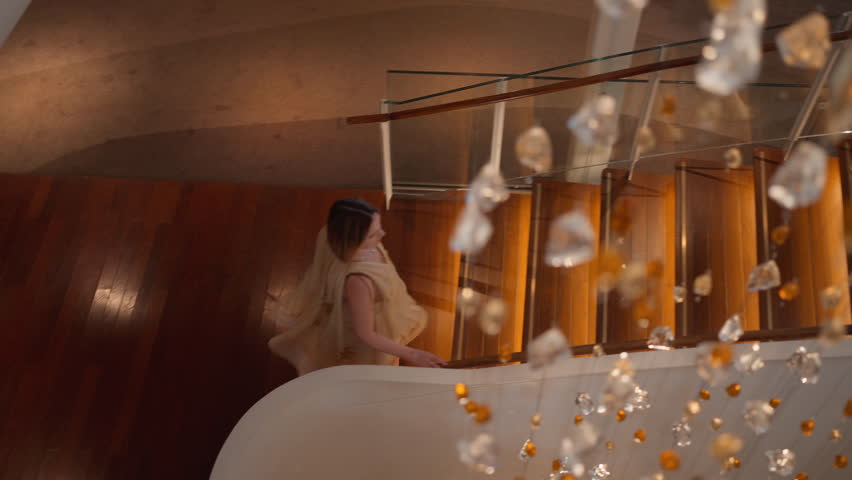 Elegant woman in flowing beige dress descends a curved wooden staircase with glass railings in a luxury hotel lobby, filmed through sparkling crystal and amber chandelier ornaments in foreground.