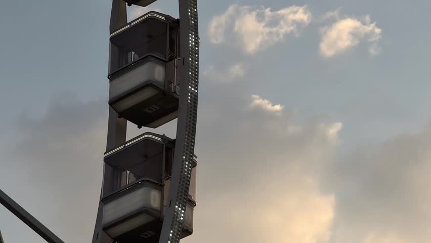 Horizontal real time video featuring a close-up view of the Budapest Ferris Wheel against a cloudy evening sky. A few passenger cabins slowly move upward while tiny LED lights shimmer across the structure. The dramatic clouds create an atmospheric urban backdrop, highlighting one of Budapest