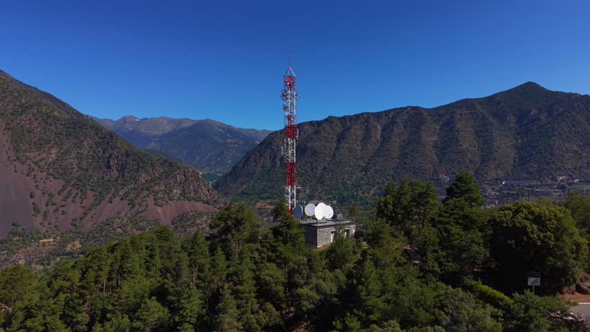 Red and white telecommunications tower with satellite dishes atop a stone building on a forested mountain ridge in Andorra, overlooking a valley town surrounded by steep slopes
