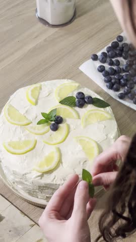 Close-up view of hands carefully placing fresh blueberries and mint sprigs onto a frosted cake adorned with lemon slices