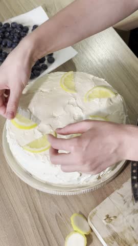 A close-up view of hands carefully placing lemon wedges and blueberries on a frosted cake, showcasing a delightful dessert preparation process