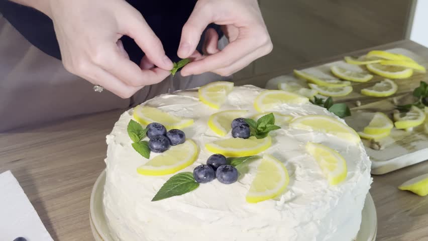 A close-up view of hands carefully placing blueberries and mint on a beautifully frosted lemon cake, ready for celebration