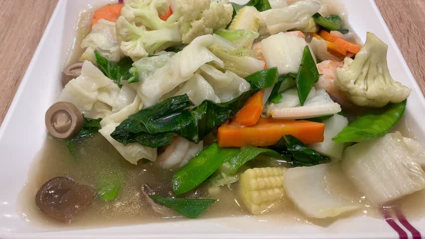 Overhead video of a healthy stir fried mixed vegetables dish with shrimp. The colorful dish features cabbage, carrots, cauliflower, baby corn and snow peas in a savory sauce served on a white plate. Ideal for cooking videos, healthy eating, and Asian food content. 