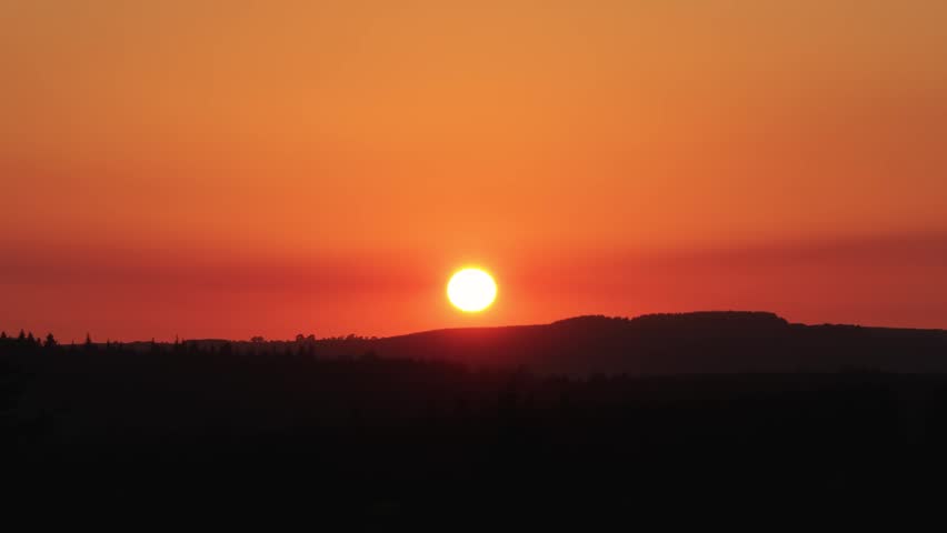 Round sun sinks behind dark Wicklow Mountains ridge as the sky glows deep orange and trees form a sharp foreground silhouette.