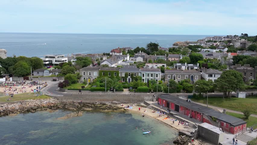 Crowds spread across the little city beach and grass park beside red bathing huts and stone harbor walls along the Irish Sea coast.
