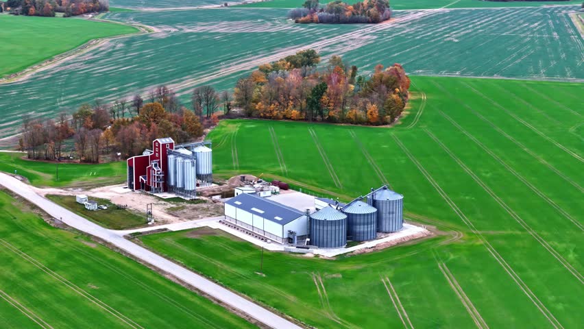 Aerial view of a farm with grain silos and a red barn structure surrounded by green agricultural fields in Riga, Latvia.
