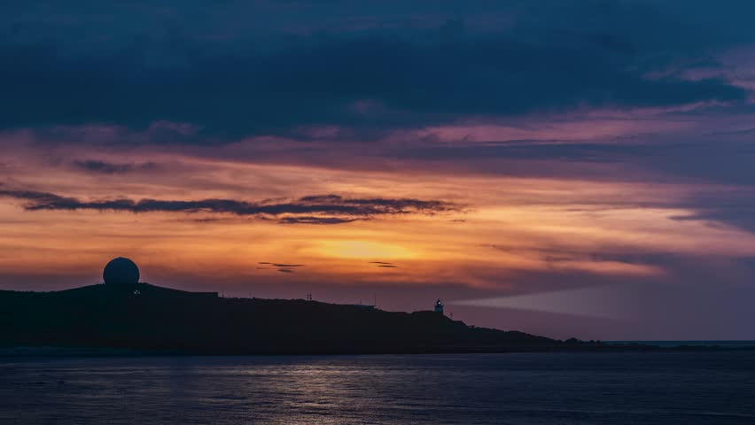 A beautiful sunset with orange and purple clouds over the coastal landscape of Shimen, Taiwan, featuring a lighthouse and radar dome silhouette.