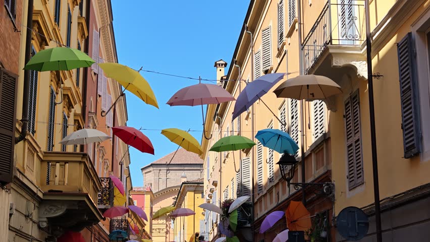 Whimsical display of vibrant umbrellas suspended above a street in the historical downtown of Modena, Italy, under a clear blue sky.