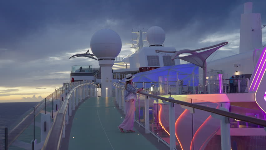 Woman in white hat stands on illuminated cruise liner deck during twilight in Caribbean Sea. Colorful neon lights glow against dramatic cloudy sky with golden sunset on horizon.