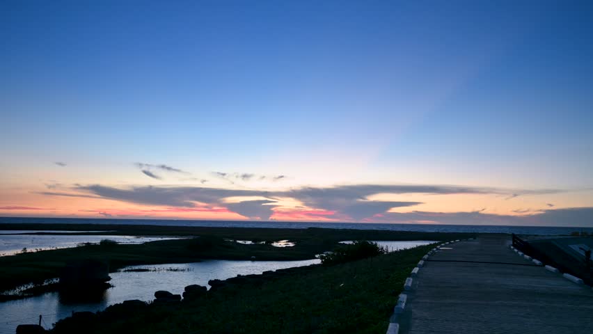 Scenic view of a coastal landscape during sunset and twilight in Miaoli, Taiwan, featuring a calm sea, wetlands, and a paved walkway.
