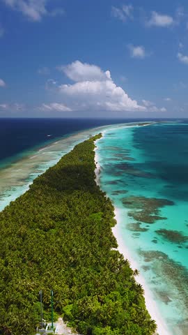 Aerial view of Dhigurah Island, Maldives, narrow palms and white sand between lagoon and ocean, bright reef bands, small boat moving on outer reef, midday light
