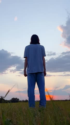 A man stands in a field, watching the sunset with hands raised. The man enjoys the open space and vibrant sky as the day ends over the grassy area.