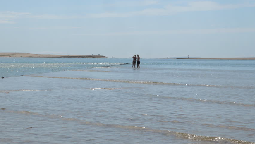 Girls Taking Selfie Standing in Shallow Lagoon Waters on Sunny Beach, Portugal. Wide shot of two girls standing in the shallow calm waters of Foz do Arelho lagoon taking a selfie, with a flat sandy horizon, sandbank, and hazy blue sky stretching into the distance along the Portuguese Silver Coast.