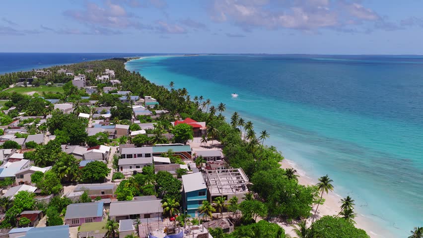 Aerial view of Dhigurah Island, Maldives, with atoll rim, white sand, coconut palms, red roofs, reef lagoon to deep ocean, speedboat wake, and gentle shore waves.