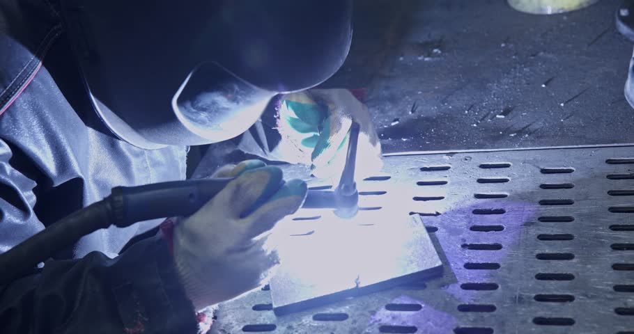 Welder joining metal plate with tig torch on perforated steel bench in industrial workshop, close handheld move documenting arc and sparks.