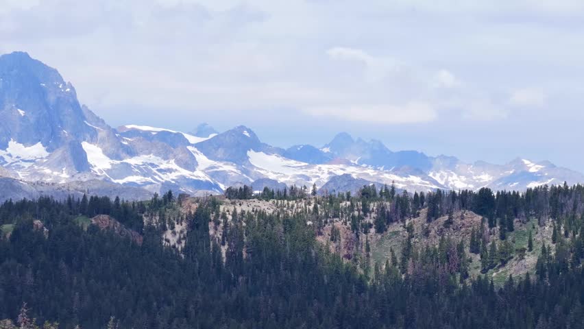 Snow-capped mountain peaks rising above dense evergreen forest ridge under cloudy sky