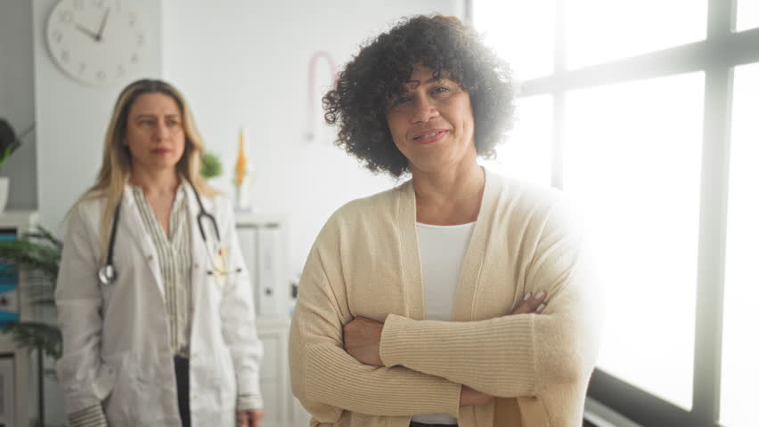 Woman patient with arms crossed and doctor wearing white coat and stethoscope placing hand on shoulder in clinic building; supportive care reassurance.
