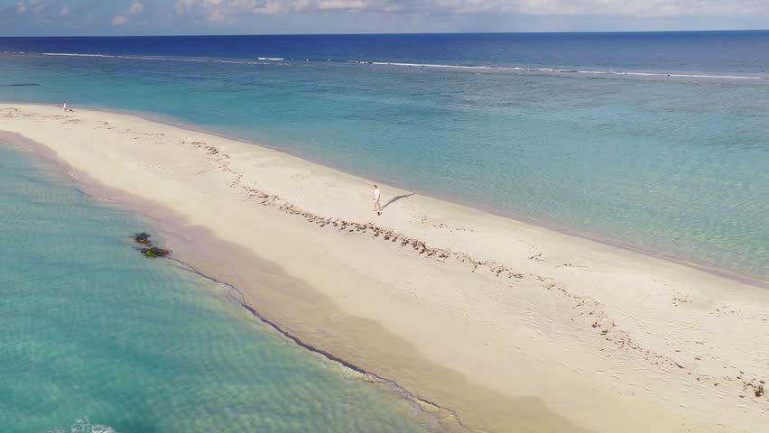 Aerial view shows a narrow sandbar and lagoon off Dhigurah, Maldives. A beautiful woman walks down the beach, waves lap both shores, a boat sits on the horizon, reefs show below.