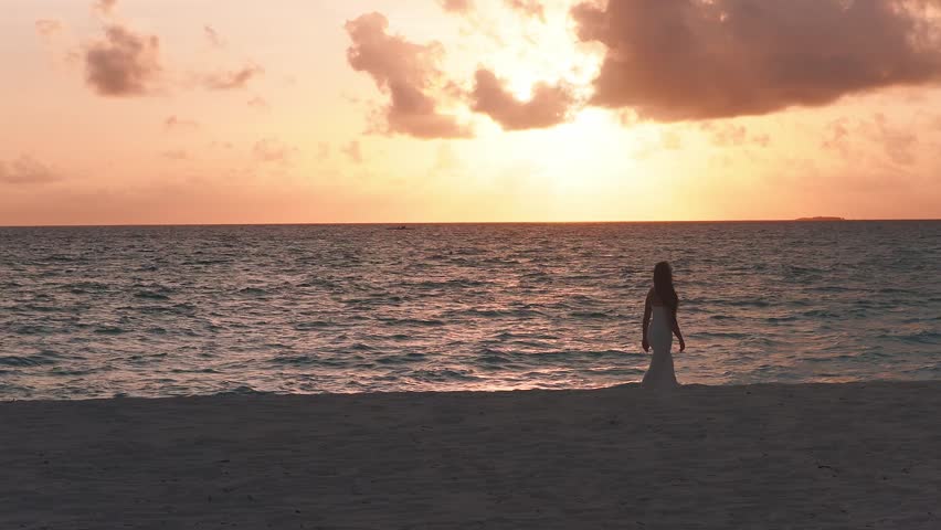Aerial view at sunset in the Maldives shows a sandy beach, a beautiful woman walking down the beach, a small jetty at left, and a distant islet. Warm sky lights clouds and rippling water.