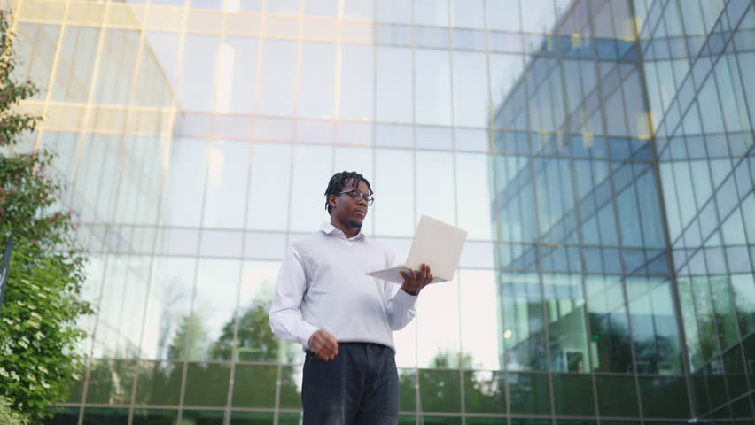 A smiling African American man stands outside a modern glass building holding a laptop and participating in a video call
