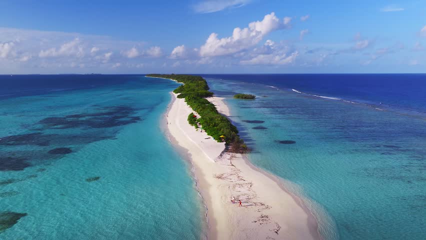 Aerial view of Dhigurah island, Maldives, shows a narrow sandbar, central green ribbon, turquoise lagoon, cobalt ocean, tiny boats, umbrellas, and walkers in bright daylight.