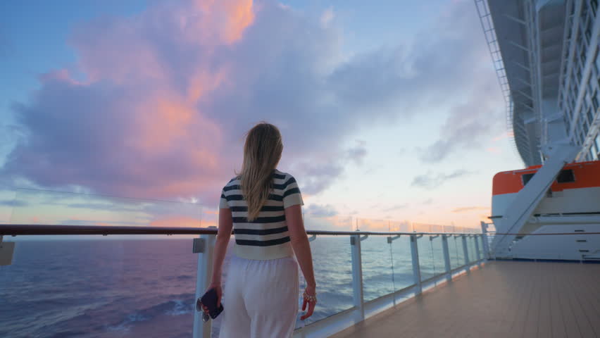 Woman in striped shirt stands on cruise liner deck in Caribbean Sea at sunset. Her hair blows in ocean breeze as pink and purple clouds fill the dramatic sky above calm waters.
