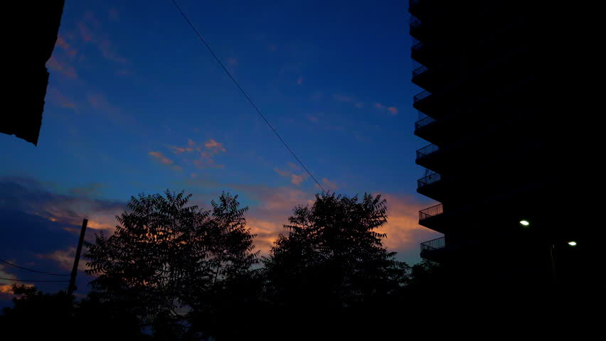 A sunset sky features vibrant colors with silhouetted trees and a high-rise building. The scene captures the tranquil beauty of dusk in an outdoor environment.