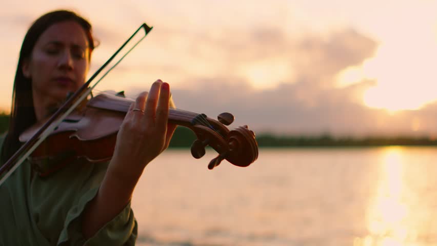 Woman violinist playing serene melody by tranquil lakeside at sunset, silhouetted against glowing sky with calm water and peaceful natural horizon