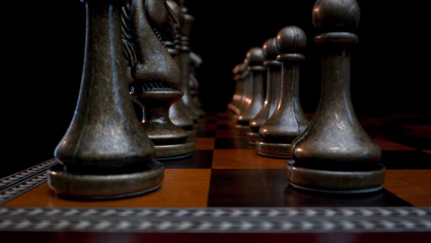 A close-up pan across a chessboard featuring polished silver and dark metallic chess pieces against a black background.