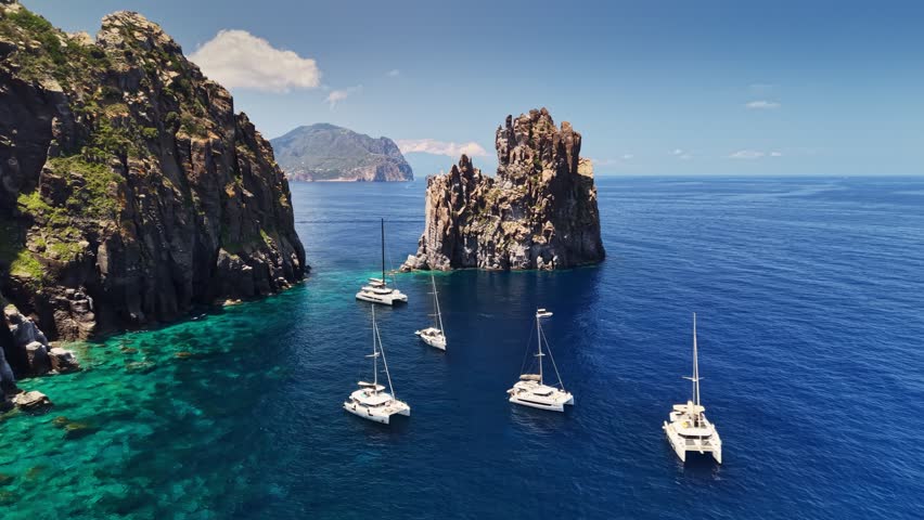 Top view of catamaran fleet floating in calm azure bay near Basiluzzo rock island, Sicily, Italy. Beautiful coastal cliffs of Aeolian islands. Vacation and yachting in the Mediterranean Sea