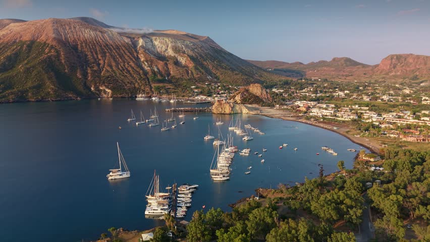 Aerial view of massive Gran Cratere volcano on Vulcano island during golden hour sunset. Sailboats lie at anchor in calm bay at mountain foot. Volcanic peak in Sicily, Aeolian archipelago, Italy