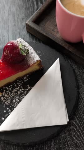 Top view of a person using a fork to eat a slice of strawberry cheesecake served with a coffee on a dark wooden table in a cafe