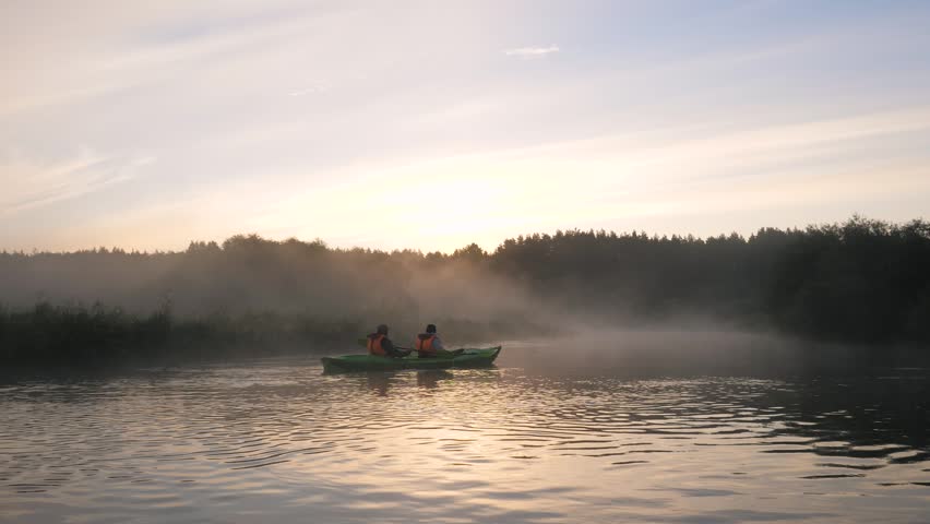 Beautiful morning landscape with mist gently rising from the surface of a tranquil river, its banks covered in lush green forest trees illuminated by the early morning sun. Aerial over river surface