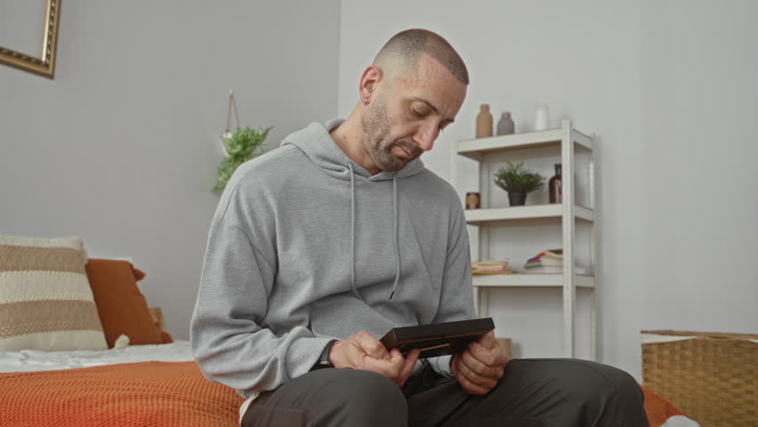 Man clutches framed photo to chest while seated on bed in a bedroom studio; grief memory reflection.