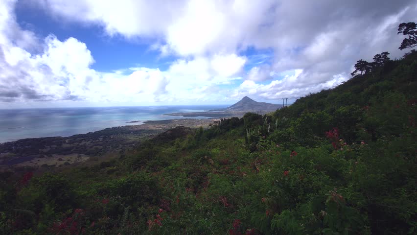 Panoramic viewpoint overlooking the lush, rolling green mountains and deep valleys of Black River Gorges National Park in Mauritius. The scenic tropical landscape features dense forest canopies under a dramatic blue sky filled with thick white clouds