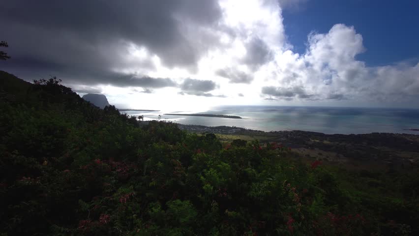 Panoramic viewpoint overlooking the lush, rolling green mountains and deep valleys of Black River Gorges National Park in Mauritius. The scenic tropical landscape features dense forest canopies under a dramatic blue sky filled with thick white clouds