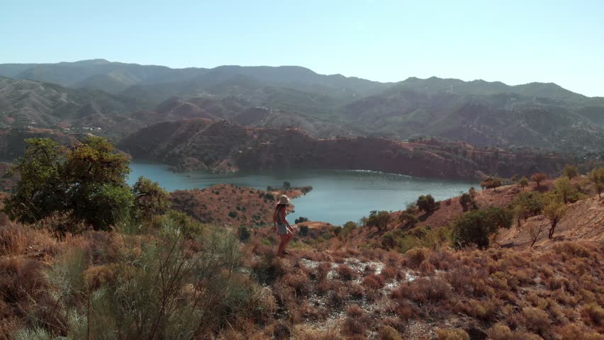 A woman in a hat and shorts walks down a dry, golden hillside covered in scrub and sparse trees, heading towards a large, tranquil reservoir. The surrounding landscape features rolling, arid mountains
