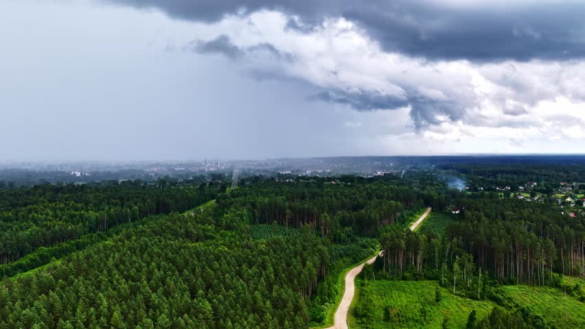 Aerial view of a winding dirt road cutting through a dense pine forest under a dramatic stormy sky near Riga, Latvia.