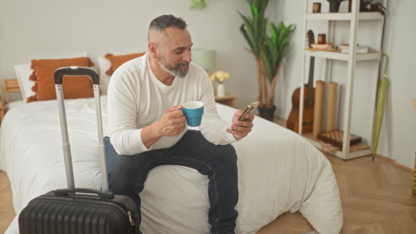 Man sitting on bed holding smartphone and blue coffee cup with suitcase beside him in building; travel planning anticipation.