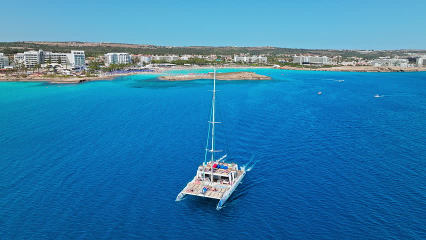 Drone view over the sea near Nissi Beach, Cyprus. A sailing boat moves through deep blue water with the beach and coastline in the distance.