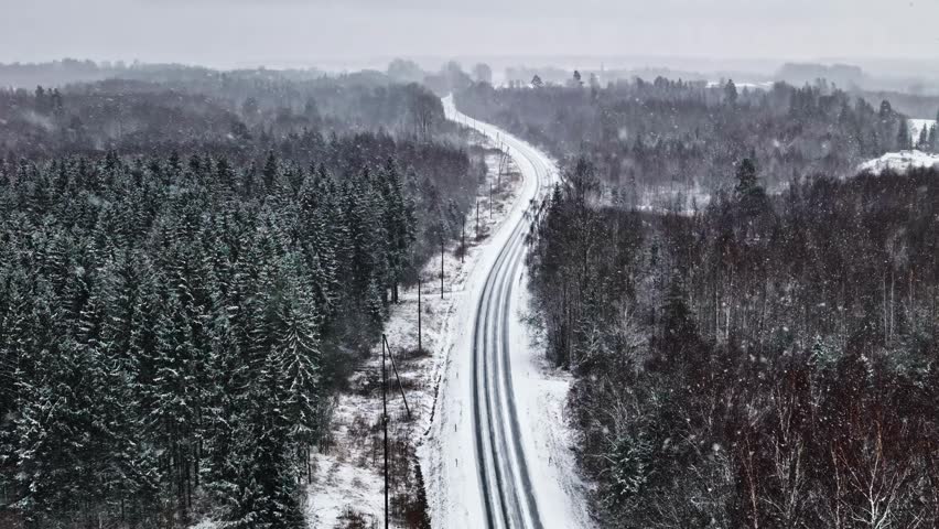 Aerial view of a snow-covered road winding through a dense pine forest during winter in Latvia.