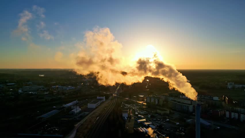 Aerial view of an industrial factory chimney emitting smoke into the golden sky at sunset in Latvia.