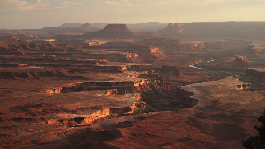 Breathtaking aerial view of Canyonlands National Park at golden hour, showcasing dramatic red rock formations, layered sandstone mesas, winding river canyon, and distant buttes under a hazy amber sky.
