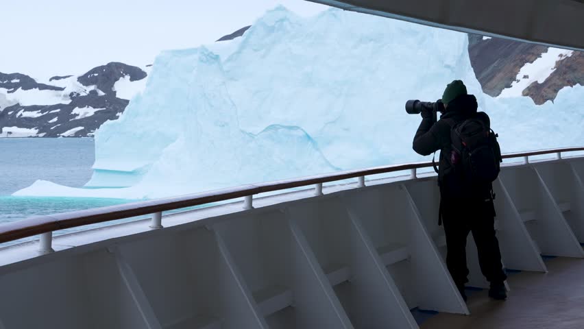 Antarctica Male Photographer on Cruise Ship Deck With Professional Camera Massive Iceberg. Exciting Adventure Travel. Nomad Black Coat, Green Hat