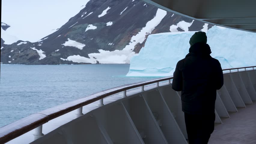 Antarctica Male Photographer Walking Cruise Ship Deck With Professional Camera Massive Iceberg. Exciting Adventure Travel. Nomad Black Coat, Green Hat