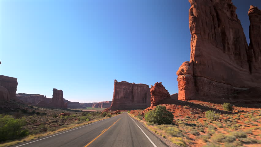 Driving along scenic highway through Arches National Park in Utah with towering red sandstone formations and buttes rising against clear blue sky. Desert vegetation lines the road.
