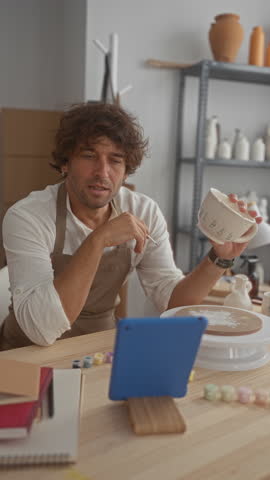 Man painting ceramic bowl with small brush in studio, holds bowl in left hand and paints delicate leaf motif; creative concentration.